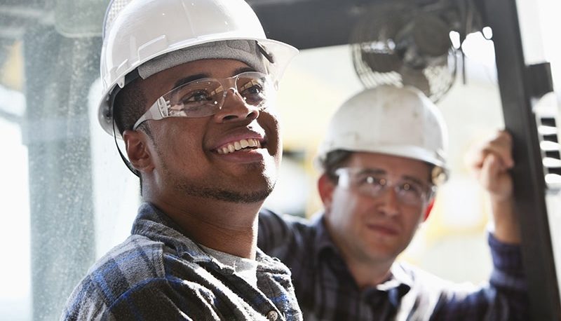 Three industrial workers are wearing hard hats and safety glasses. The African American in the foreground of the picture is holding a document. There is a piece of heavy duty equipment shown in the background. One of the workers is sitting on the heavy duty equipment.